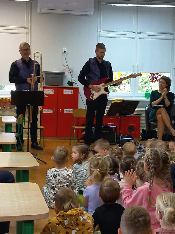 Pani siedzi na krześle. jeden pan stoi i gra na gitarze elektrycznej, drugi pan trzyma w ręku puzon.