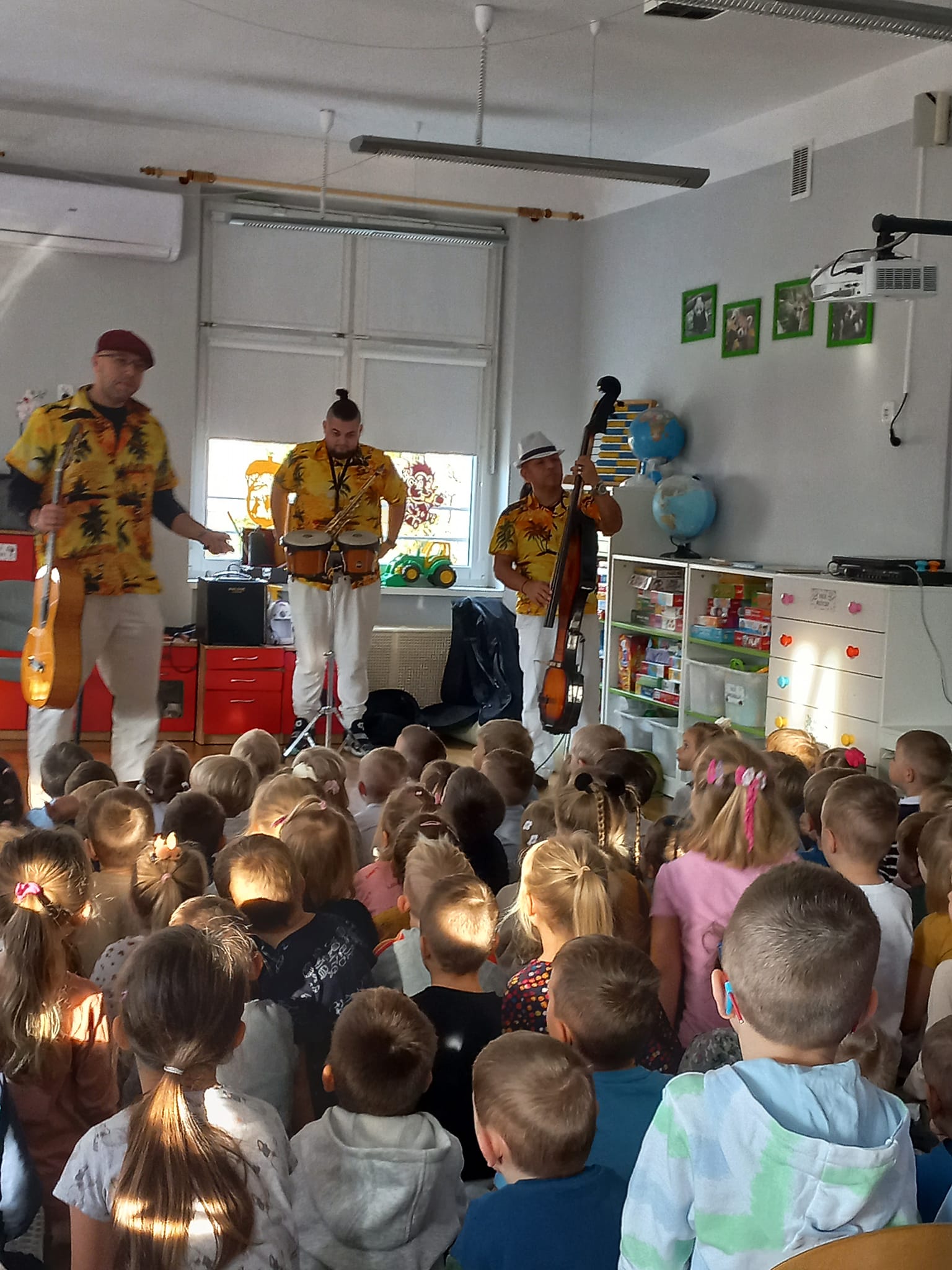 Trzech pan&oacute;w gra na instrumentach: saksofon, gitara, bongosy. Dzieci siedzą na dywanie.
