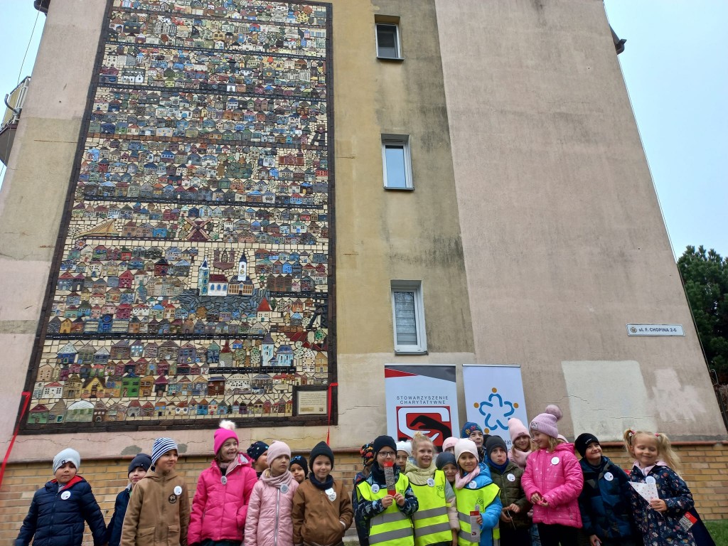 Grupa dzieci stoi na tle ceramicznej mozaiki przymocowanej na ścianie bloku.