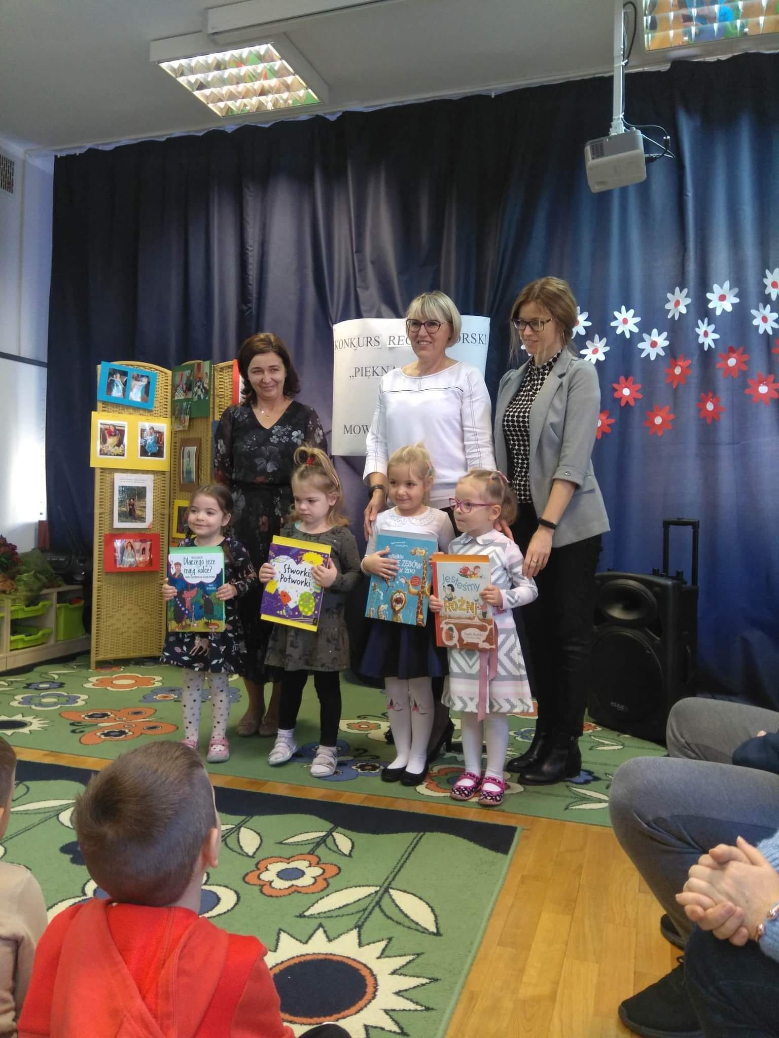 Cztery dziewczynki stoją trzymając książki i dyplomy za zajęte miejsca w konkursie. Za dziewczynkami stoją trzy panie.