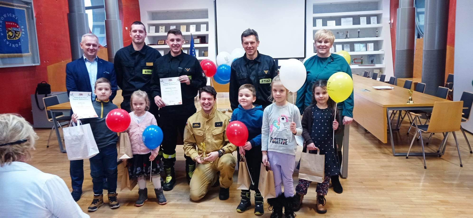 Grupa dzieci ze strażakami. Wręczenie nagr&oacute;d za konkurs.