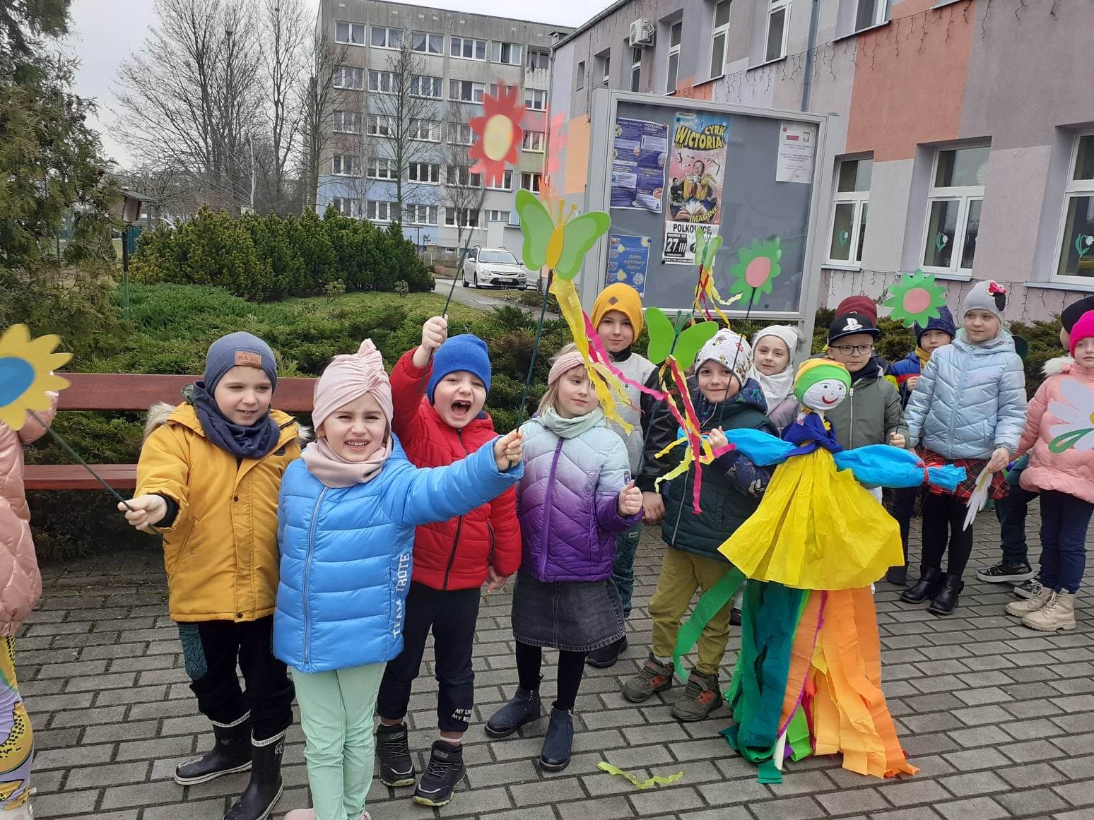 Dzieci stoją przed przedszkolem i witają wiosnę machając korowymi kwiatami