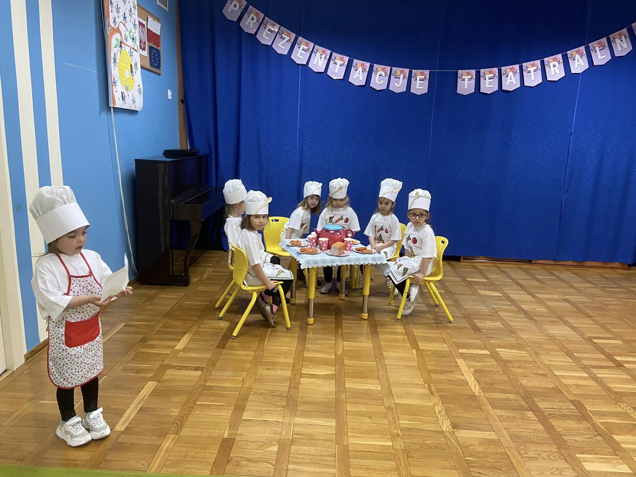 Sześć dziewczynek przebranych za kucharki siedzi przy stoliku. Jedna dziewczynka stoi 