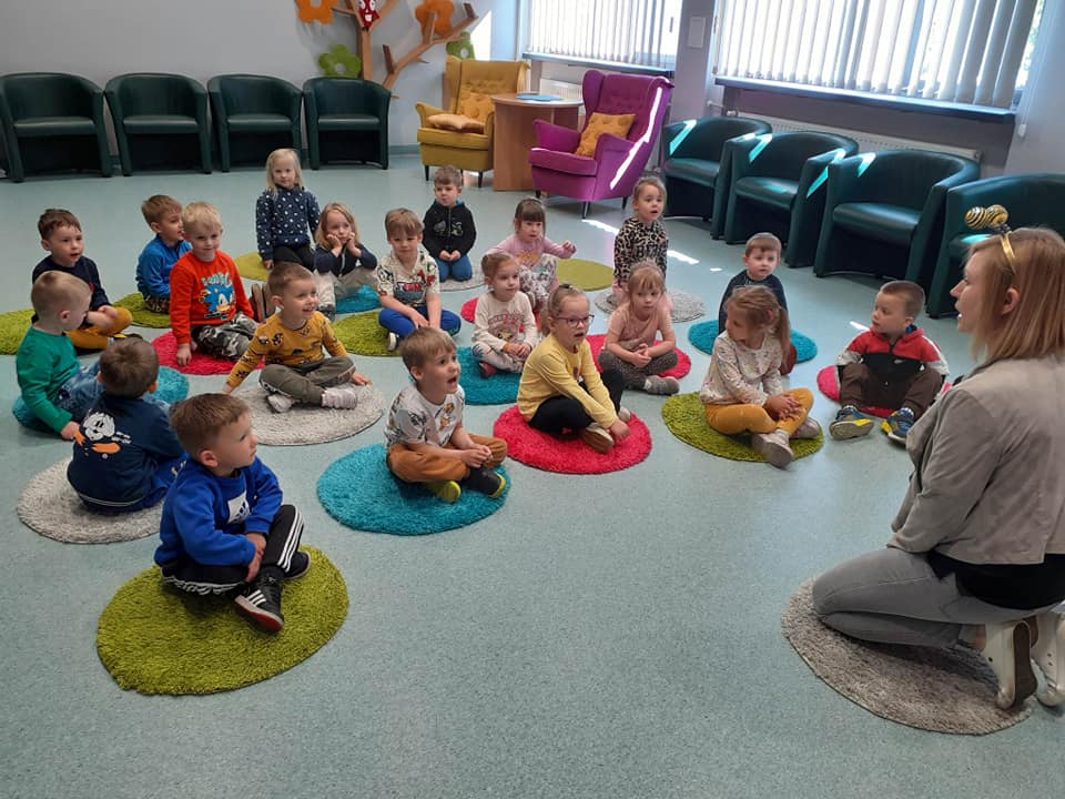 Dzieci siedzą na małych dywanikach na zajęciach w bibliotece.