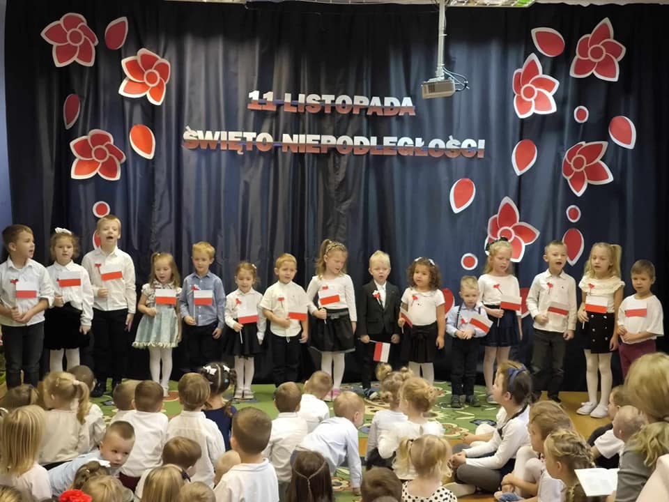 Dzieci ubrane na galowo trzymają w ręku flagę Polski i śpiewają.