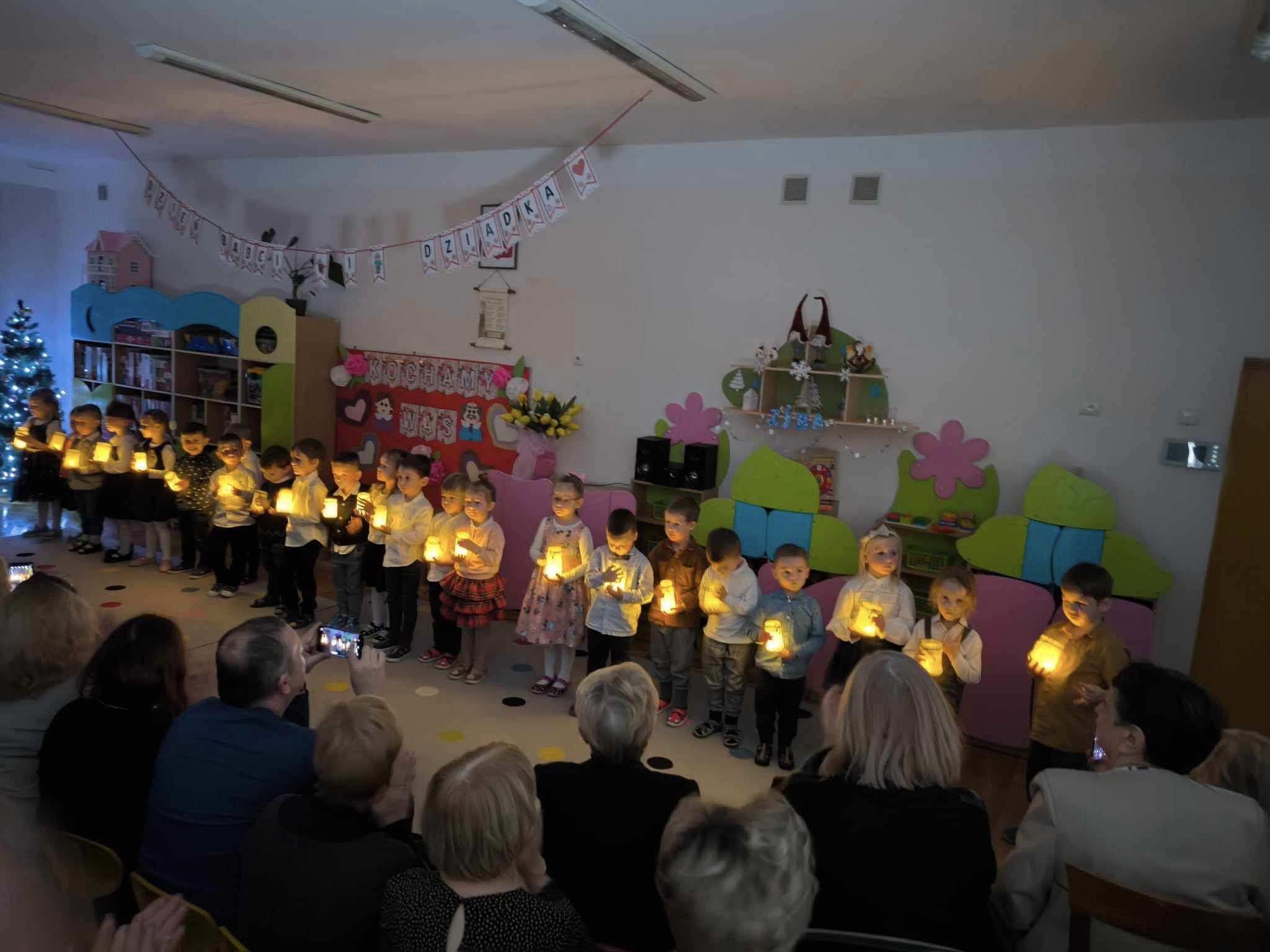Dzieci stoją w p&oacute;łkolu trzymając w ręku zapalone lampki.