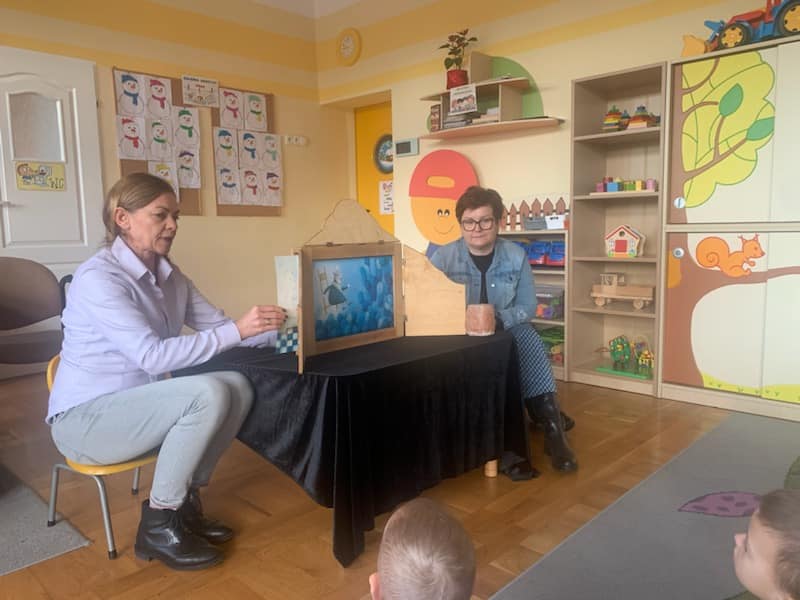 Dwie panie siedzą na krzesełkach i przedstawiają teatrzyk Kamishibai.