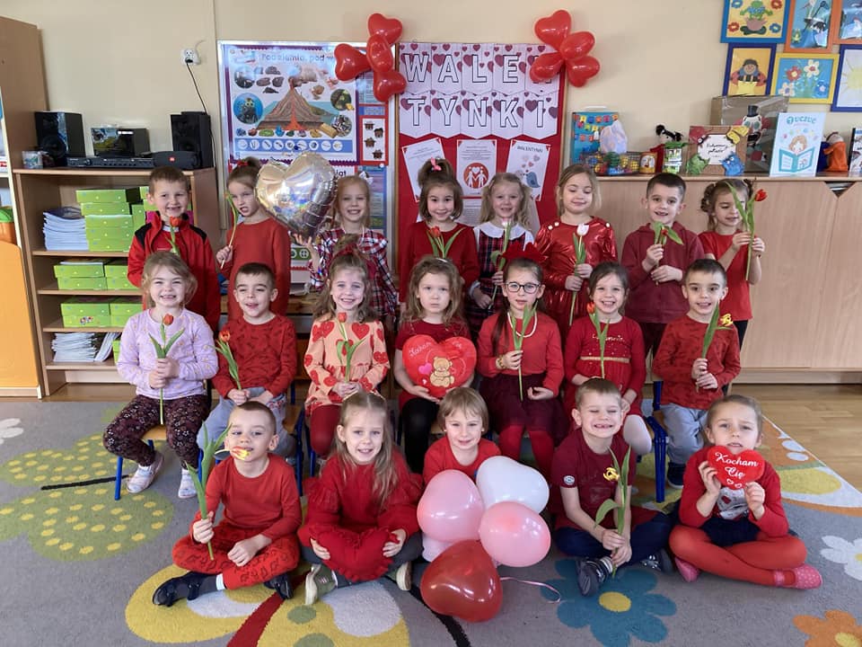 Dzieci ubrane na czerwono stoją w grupie trzymając w ręku kwiatka lub serce.