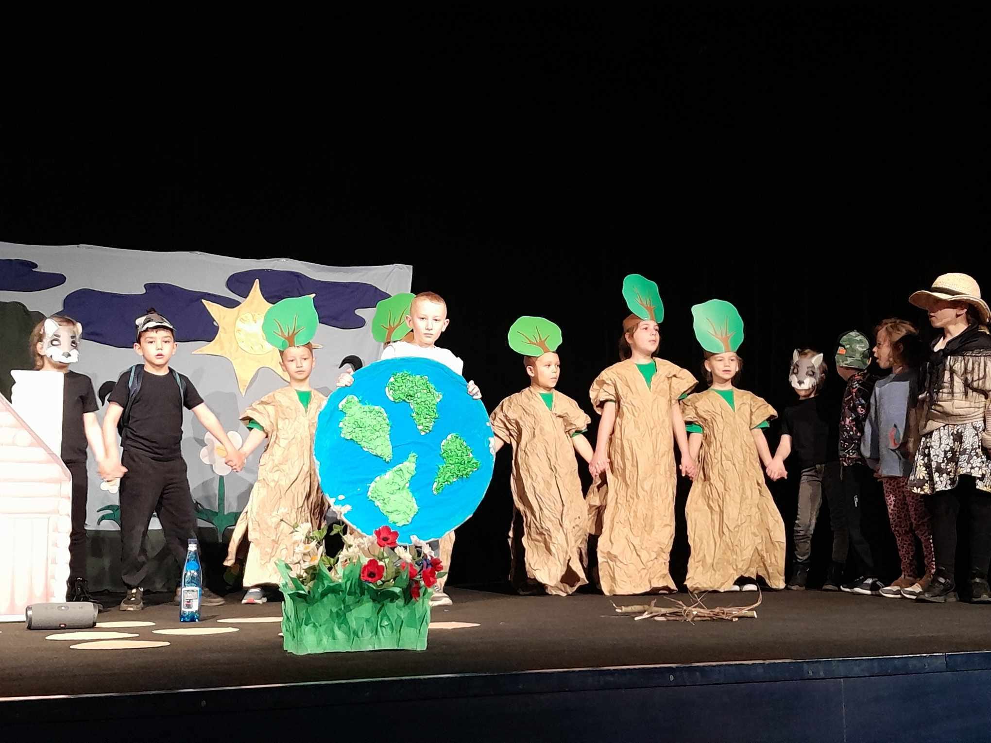 Dzieci przebrane za drzewa i zwierzęta stoją na scenie.