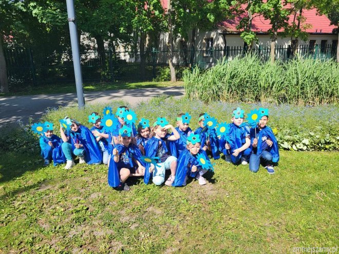 Dzieci ubrane w niebieskie pelerynki z opaskami kwiatowymi  na głowie kucają na trawie.