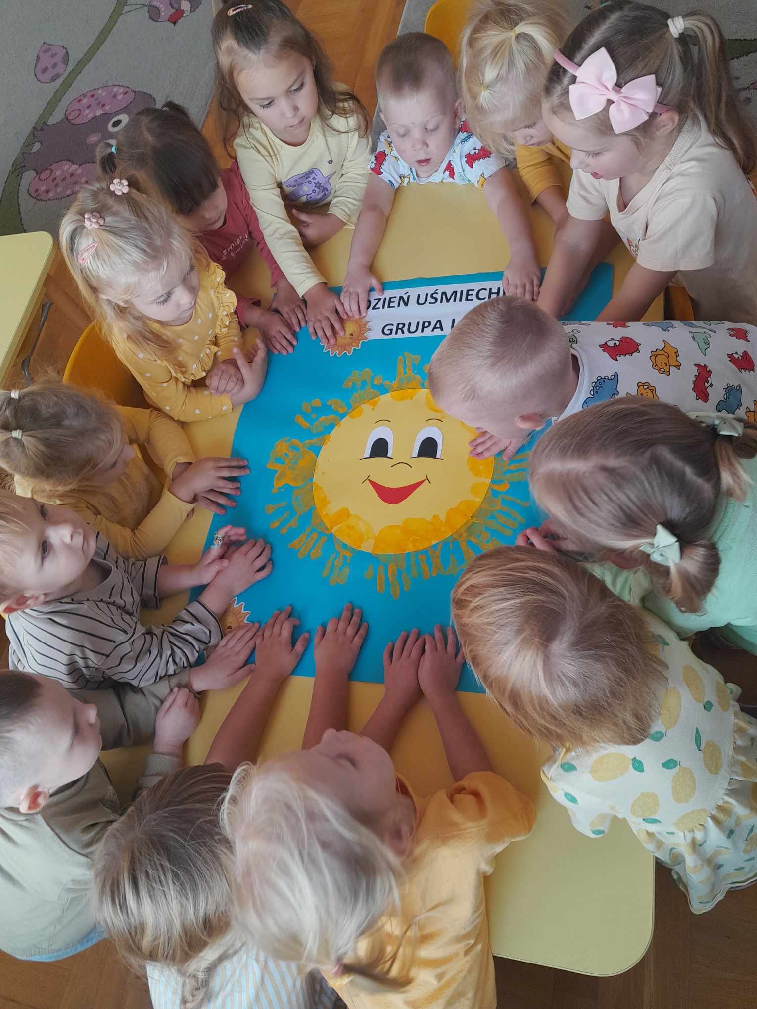 Dzieci ubrane na ż&oacute;łto stoją wok&oacute;ł plakatu ze słońcem i odciśniętymi rękami.