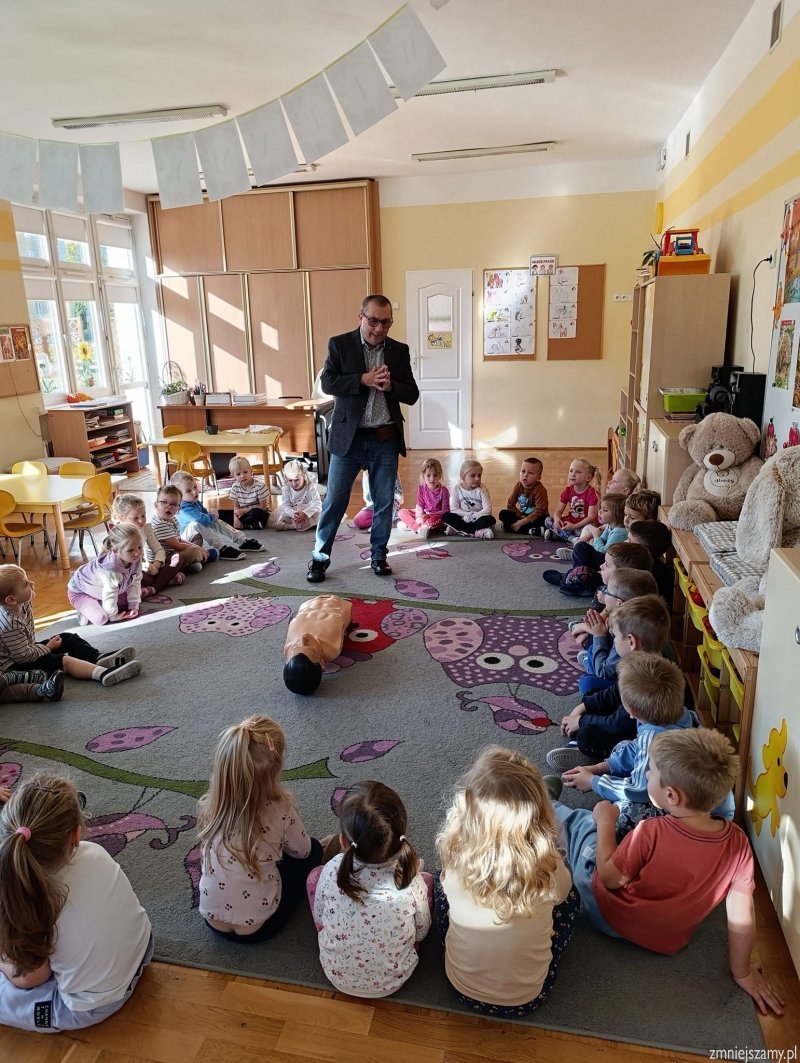 Na dywanie leży fantom, dzieci siedzą w kole słuchając pana.