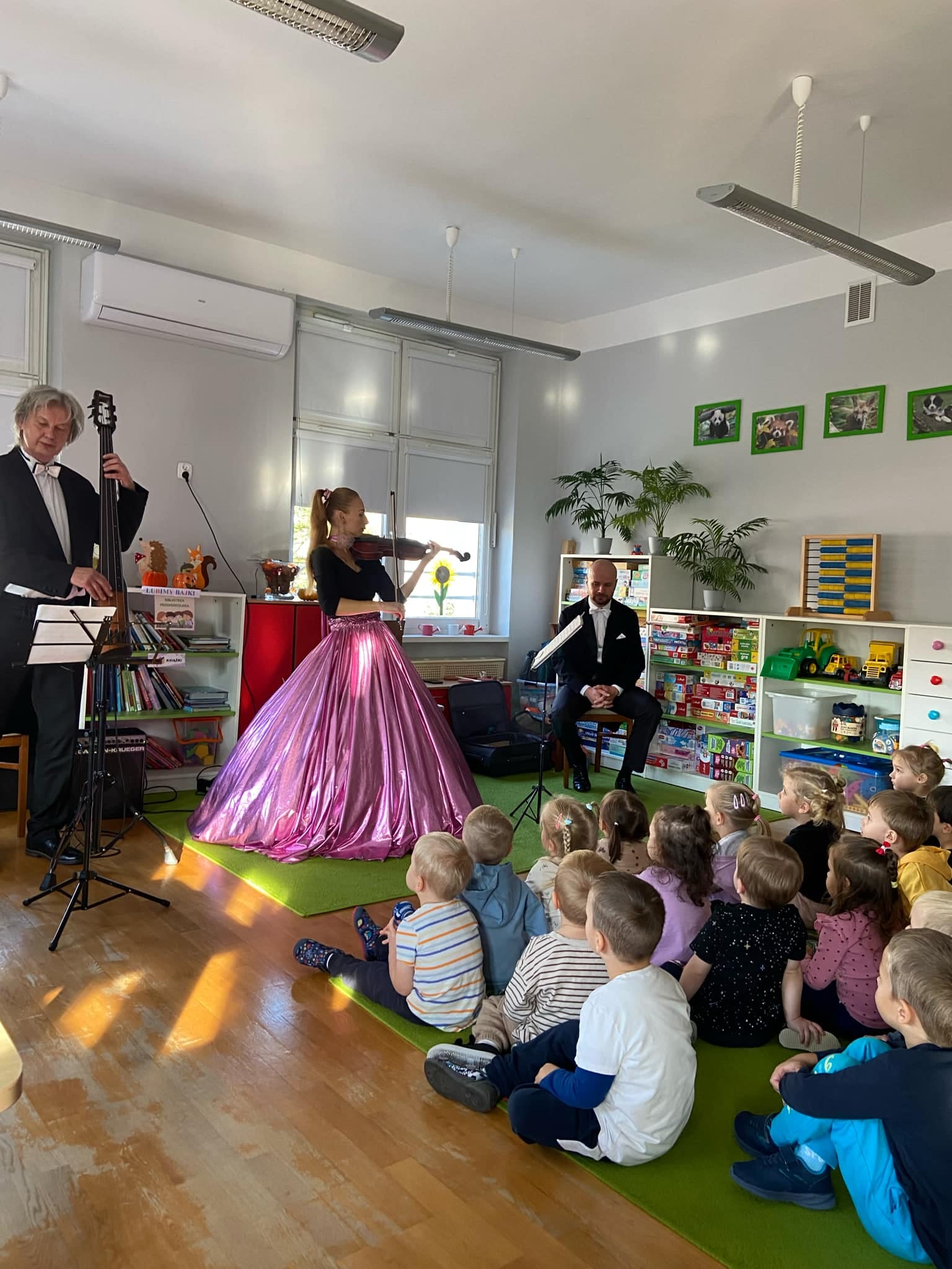 Troje muzyk&oacute;w gra na instrumentach muzycznych dzieci siedzą na dywanie i słuchają.