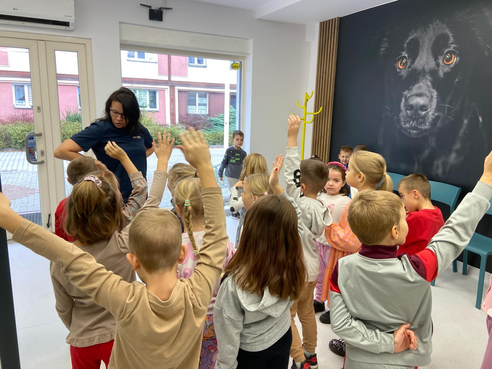 Dzieci  stoją w grupie słuchając pani weterynarz. Na ścianie obraz psa.