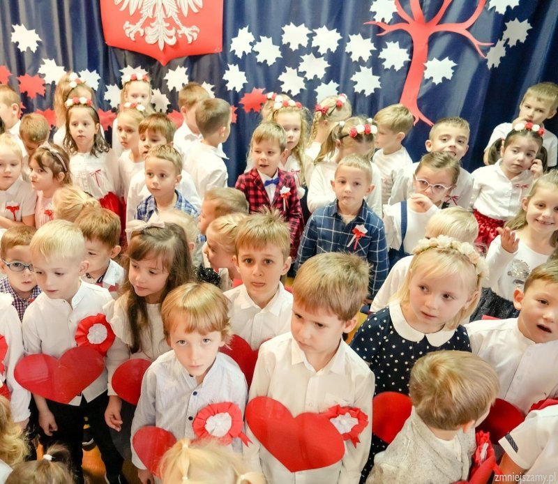 Dzieci ubrane na galowo trzymają w ręku czerwone serca z papieru i śpiewają.