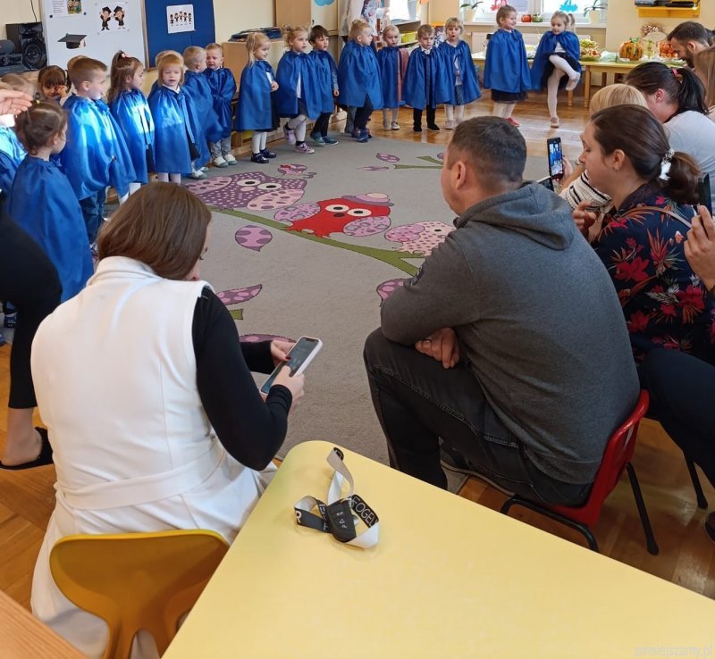 Dzieci w niebieskich pelerynkach stoją w p&oacute;łkolu. Rodzice oglądają występ.