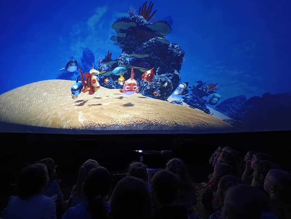 Dzieci siedzą pod kopułą i oglądają film o rybkach.