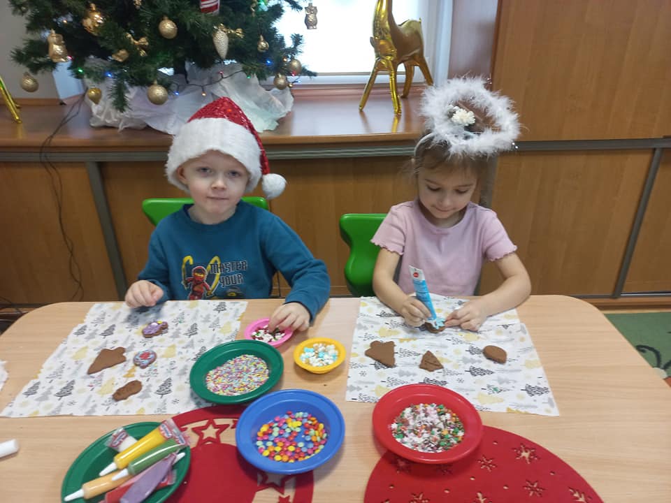 Chłopiec w mikołajkowej czapce i dziewczynka w aureoli siedzą przy stole i dekorują świąteczne pierniki.