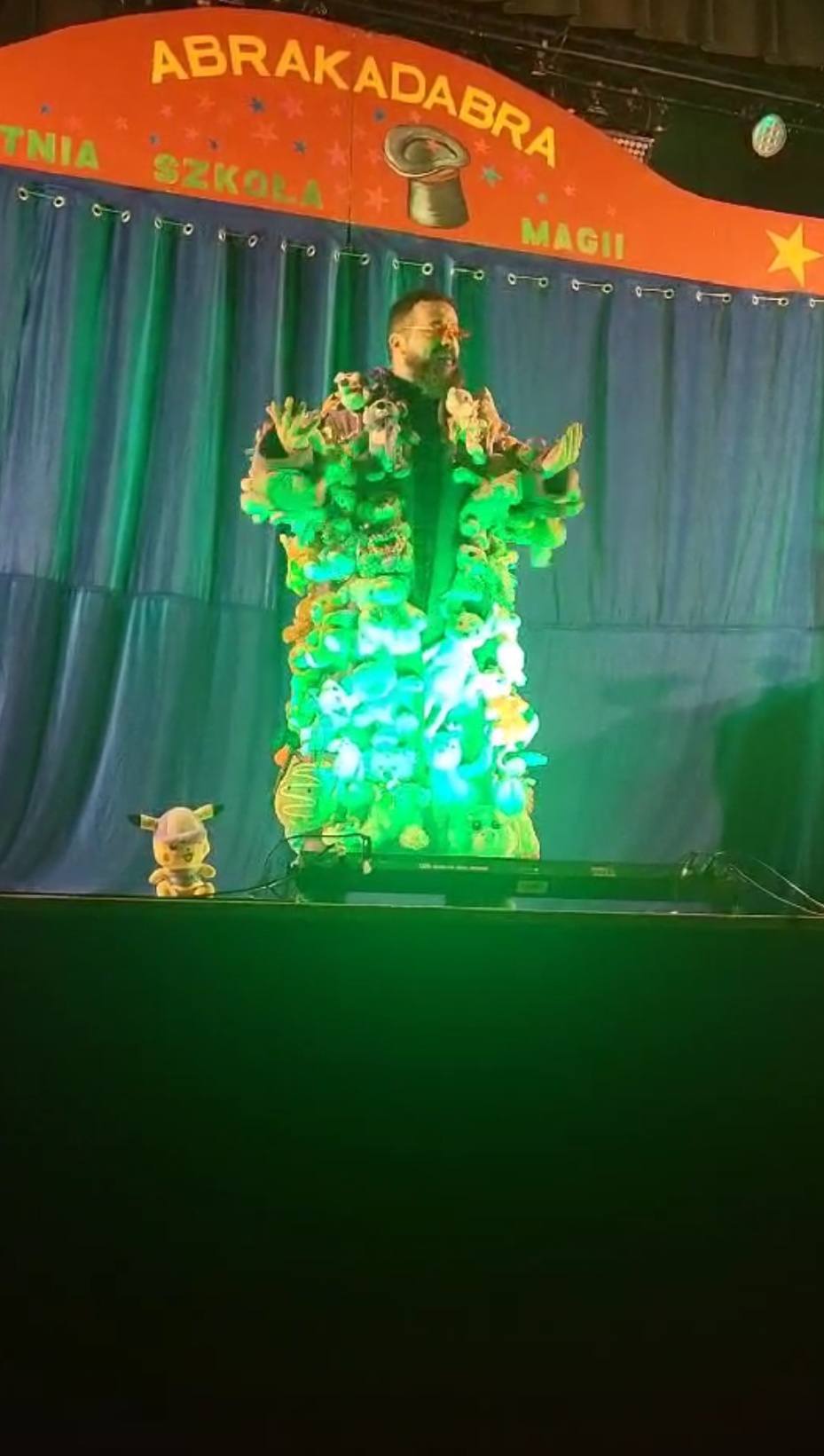 Na scenie stoi czarodziej ubrany w płaszcz ozdobiony maskotkami.