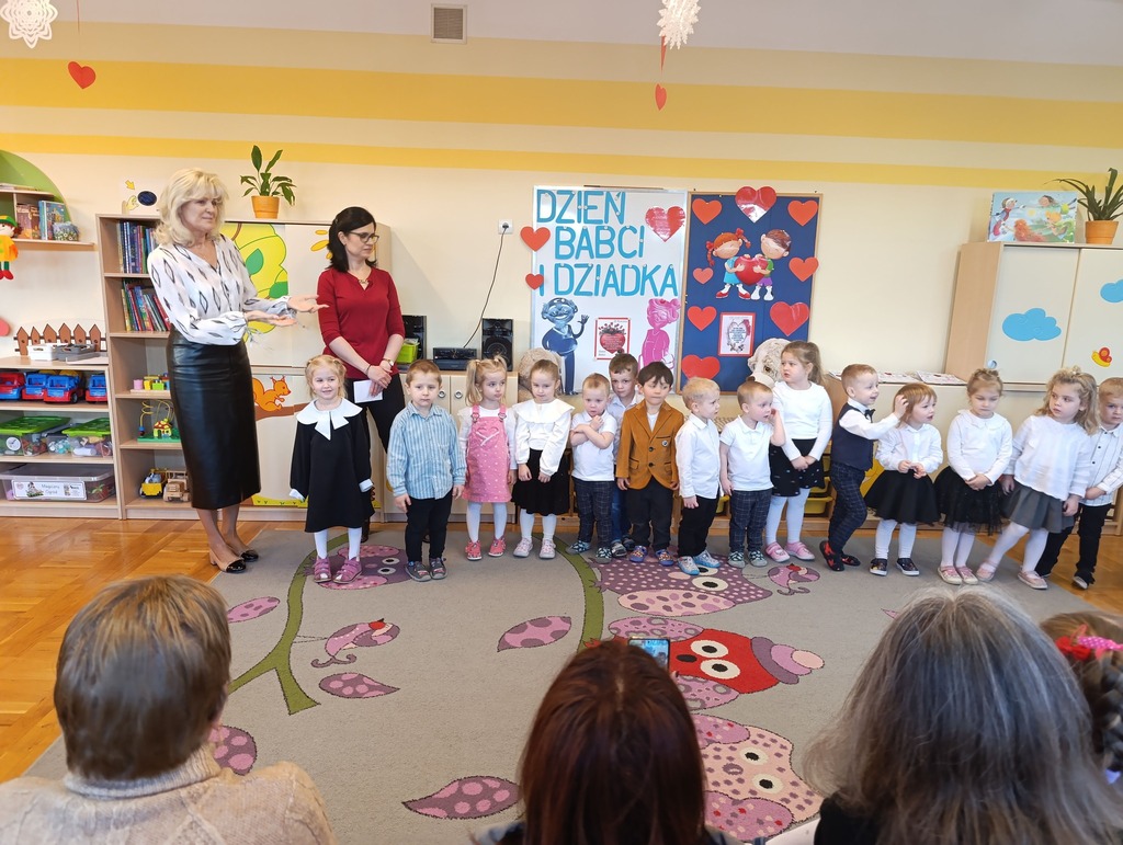 Dzieci ubrane na galowo stoją w p&oacute;łkolu razem z dwoma paniami.