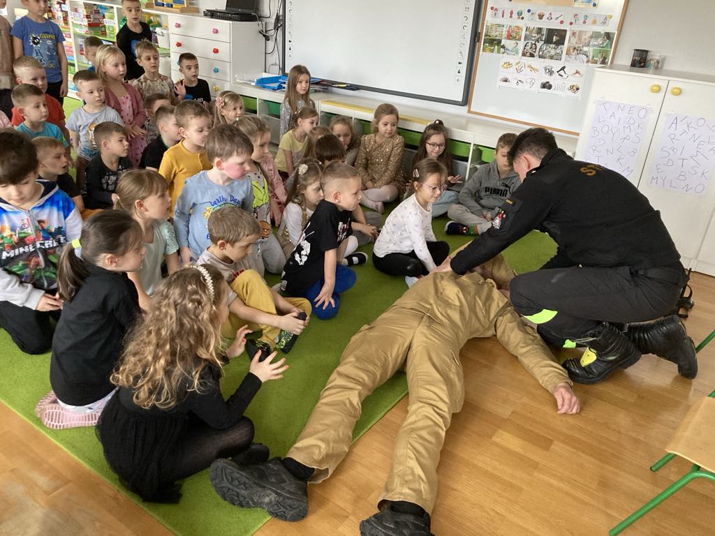 Dzieci siędza na dywanie dw&oacute;ch strażak&oacute;w pokazuje jak prawidłowo udzielać pierwszej pomocy.