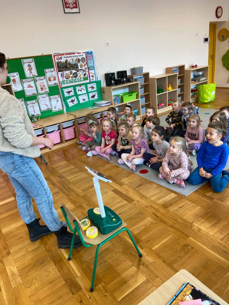 Dzieci siedzą w grupie na dywanie i słuchają pani, kt&oacute;ra opowiada o  energii odnawialnej.j.i 