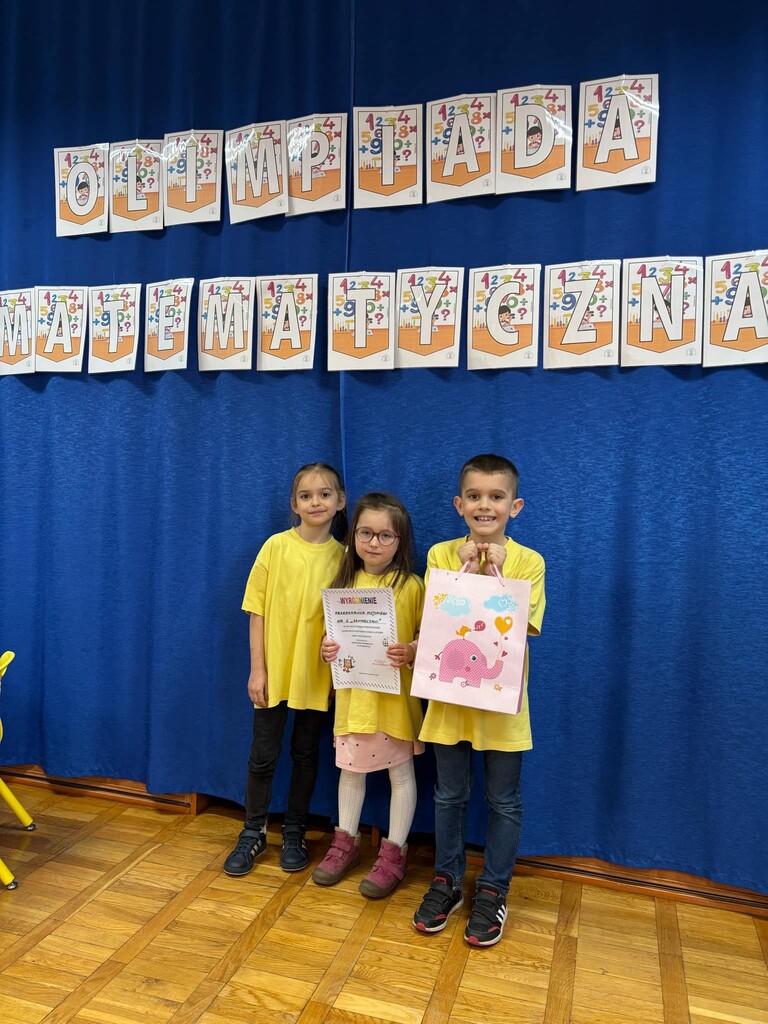 Troje dzieci stoi z dyplomem na tle napisu olimpiada matematyczna.