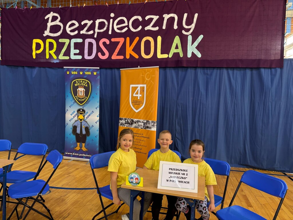 Dzieci ubrane w ż&oacute;łte koszulki siedzą przy stoliku na tle napisu bezpieczny przedszkolak.