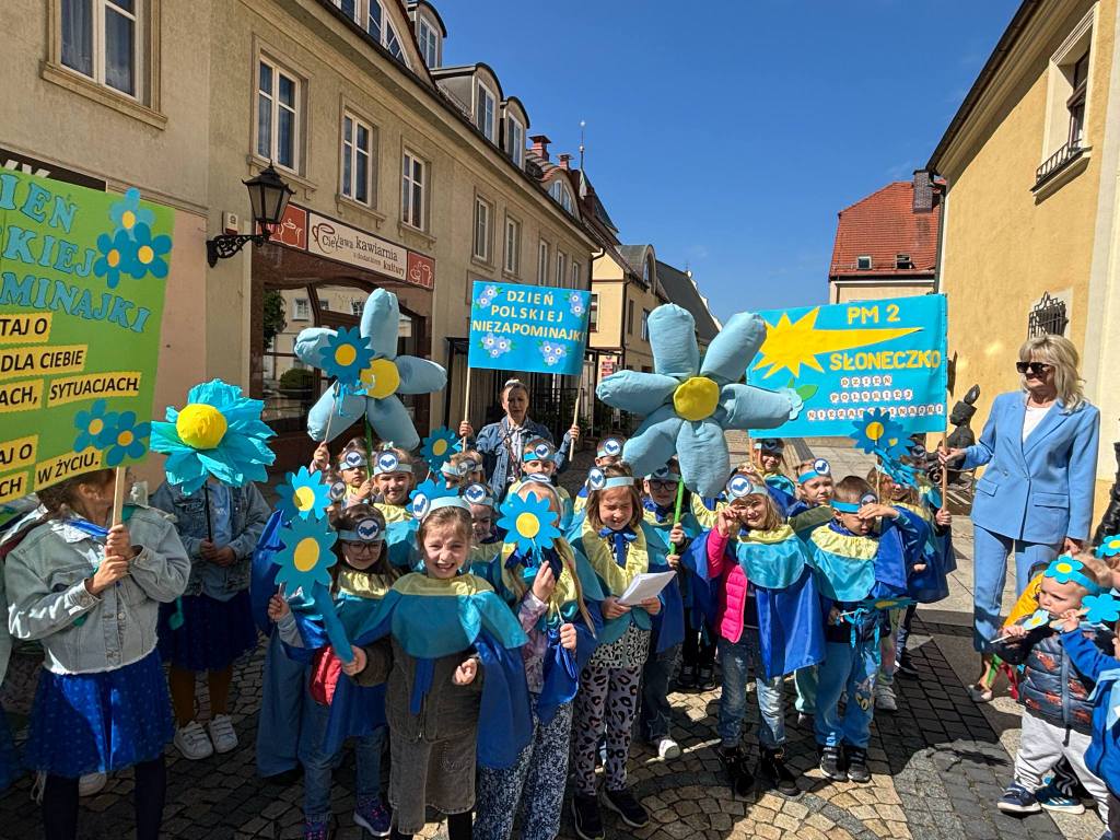 Dzieci ubrane na niebisko trzymają w ręku niebieskie kwiaty oraz plakaty.