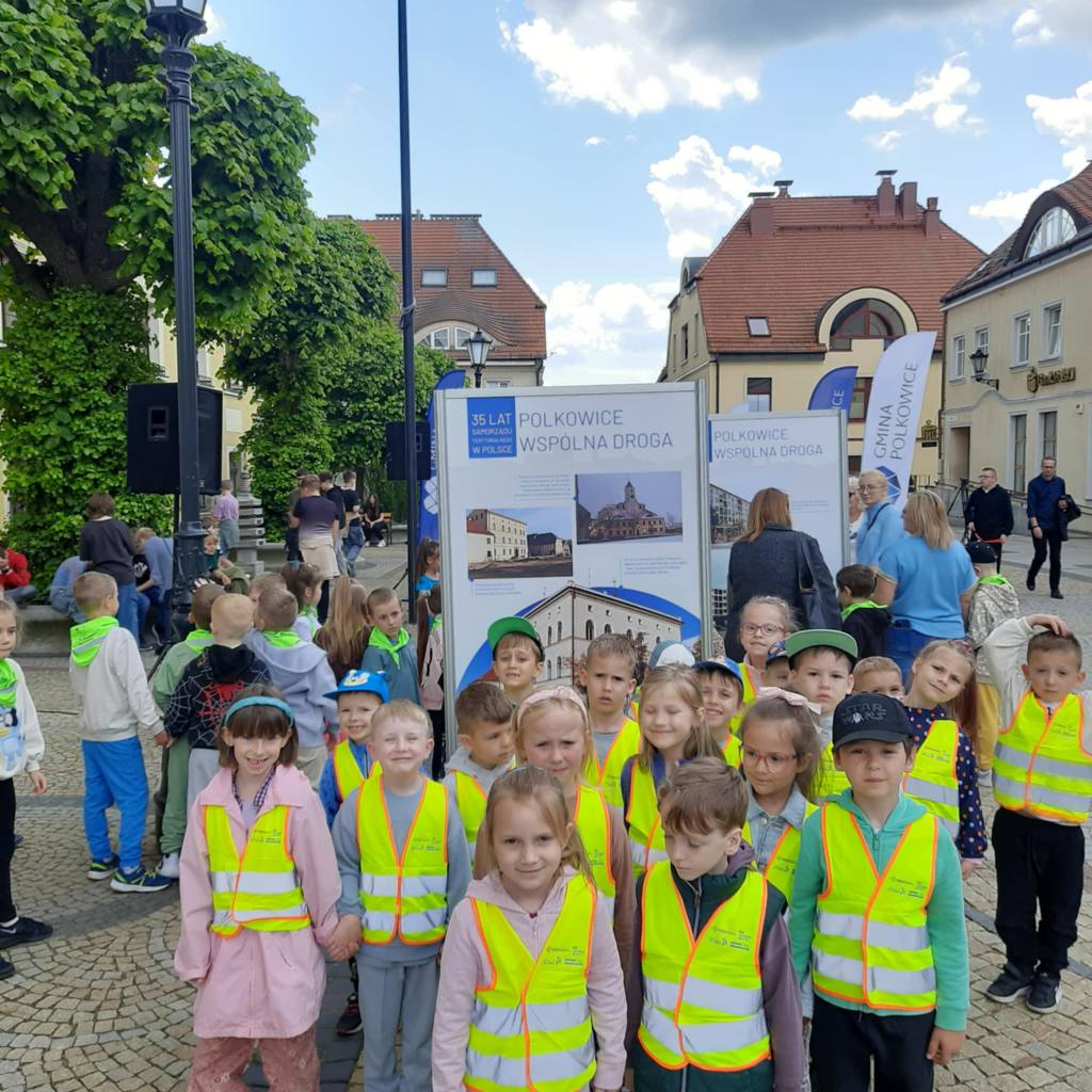 Dzieci stoja w grupie w kamizelkach odblaskowych na tle wystawy przedstawiającej Polkowice.
