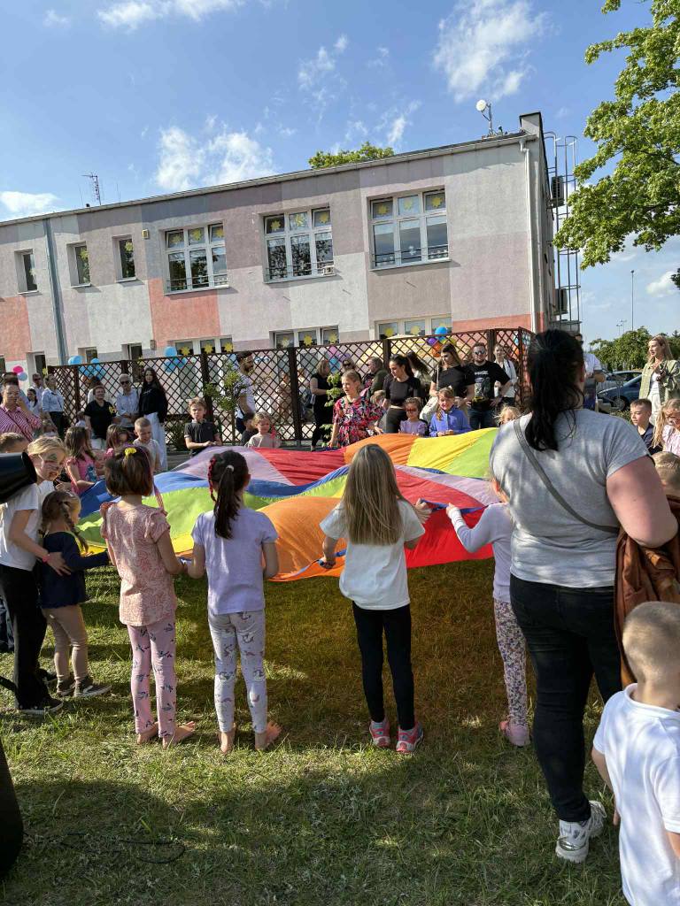 Dzieci wraz z rodzicami trzymają kolorową chustę animacyjna w ogrodzie przedszkolnym.