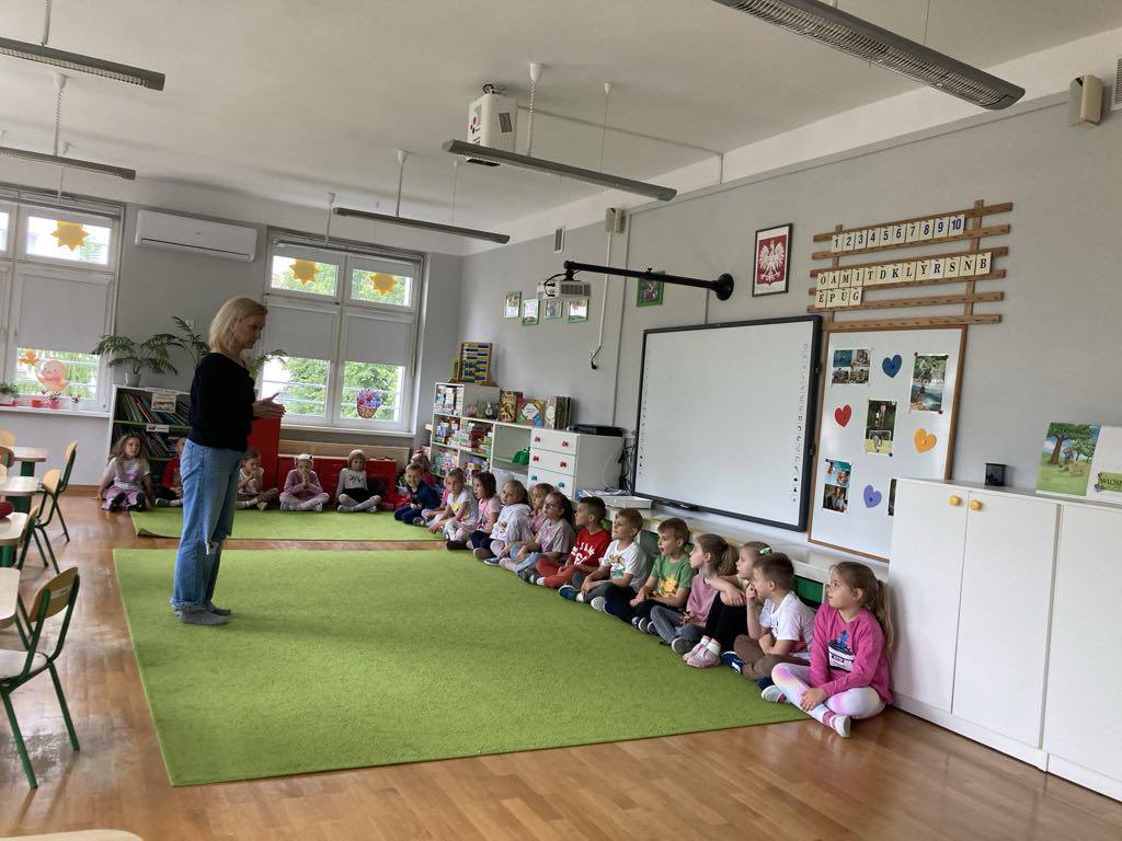 Dzieci siedzą w p&oacute;łkolu na dywanie razem z aktorką.