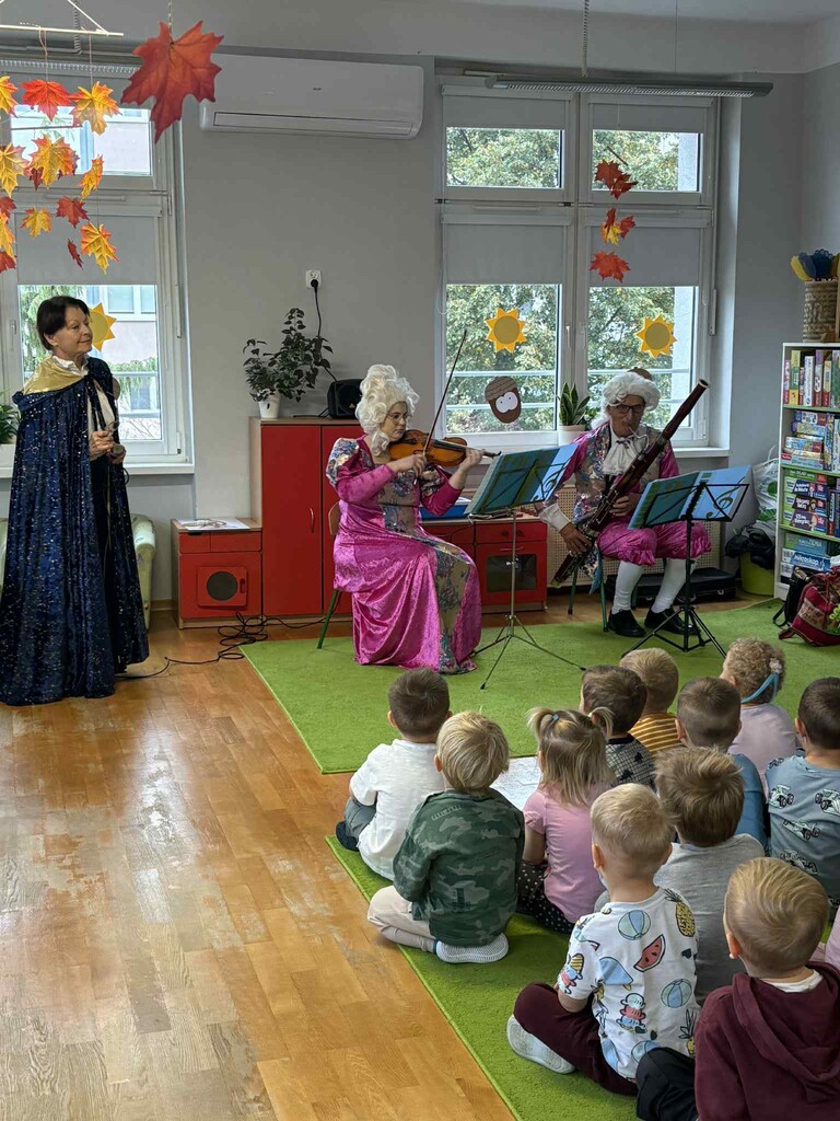 Dzieci siedząc na dywanie słuchają koncertu w  wykonaniu trzech muzyk&oacute;w