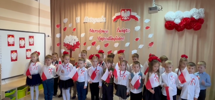 Dzieci ubrane na galowo stoją w grupie trzymając w ręku flagi Polski