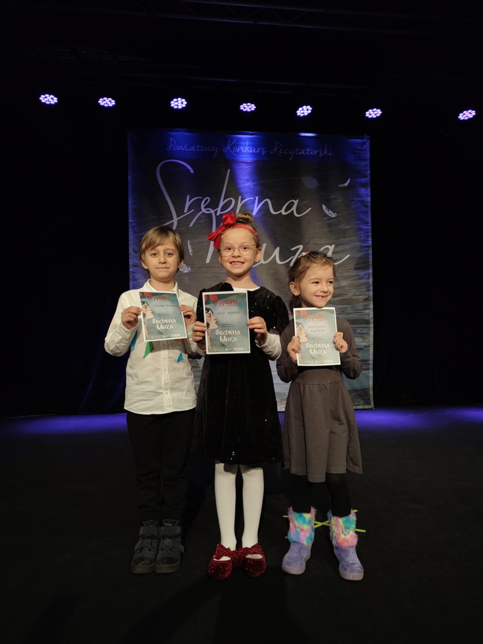 Troje dzieci stoi na scenie z dyplomami.