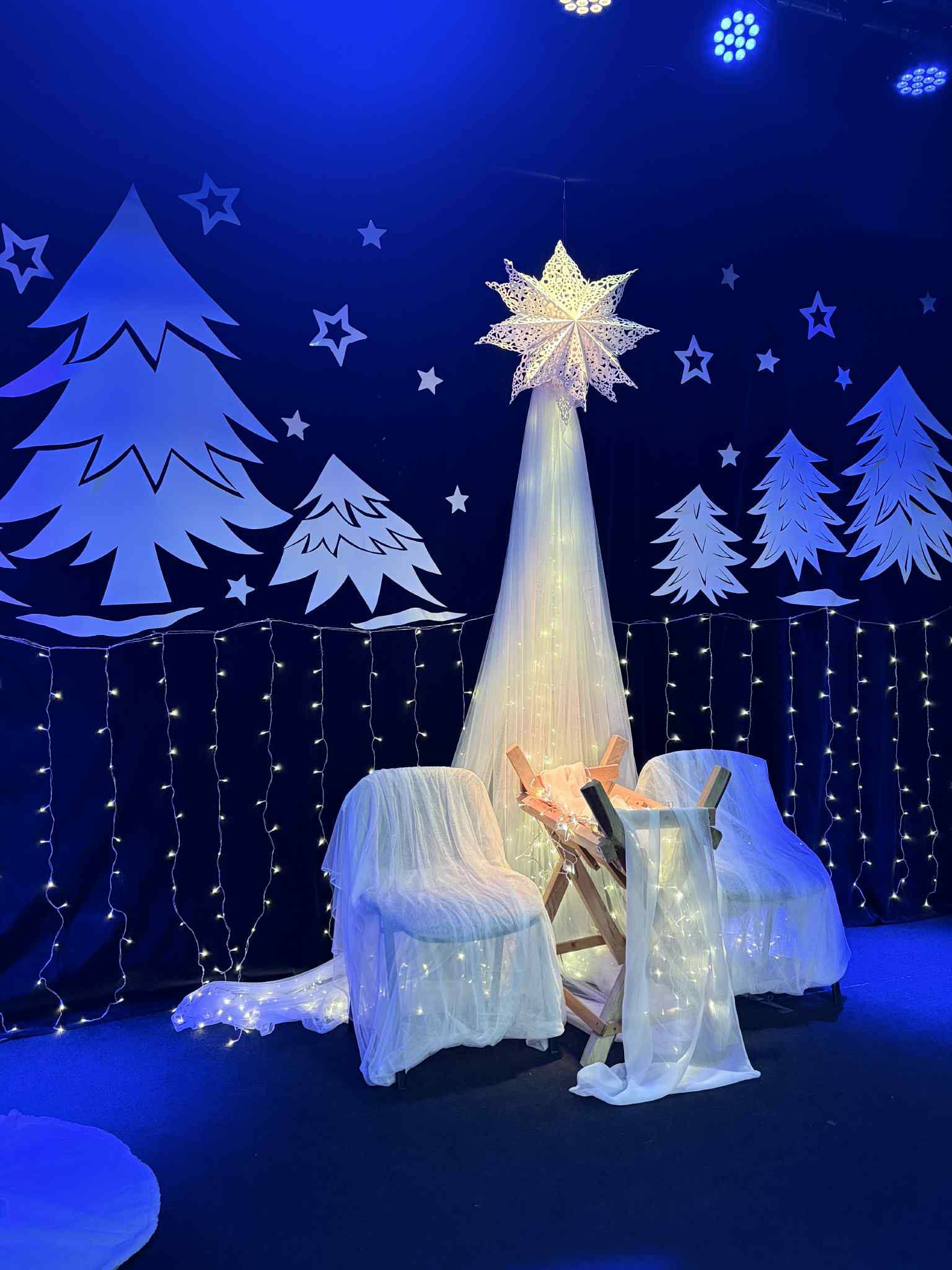 Na scenie białe choinki przyczepione do kotary, stoi żłobek i gwiazda betlejemska.