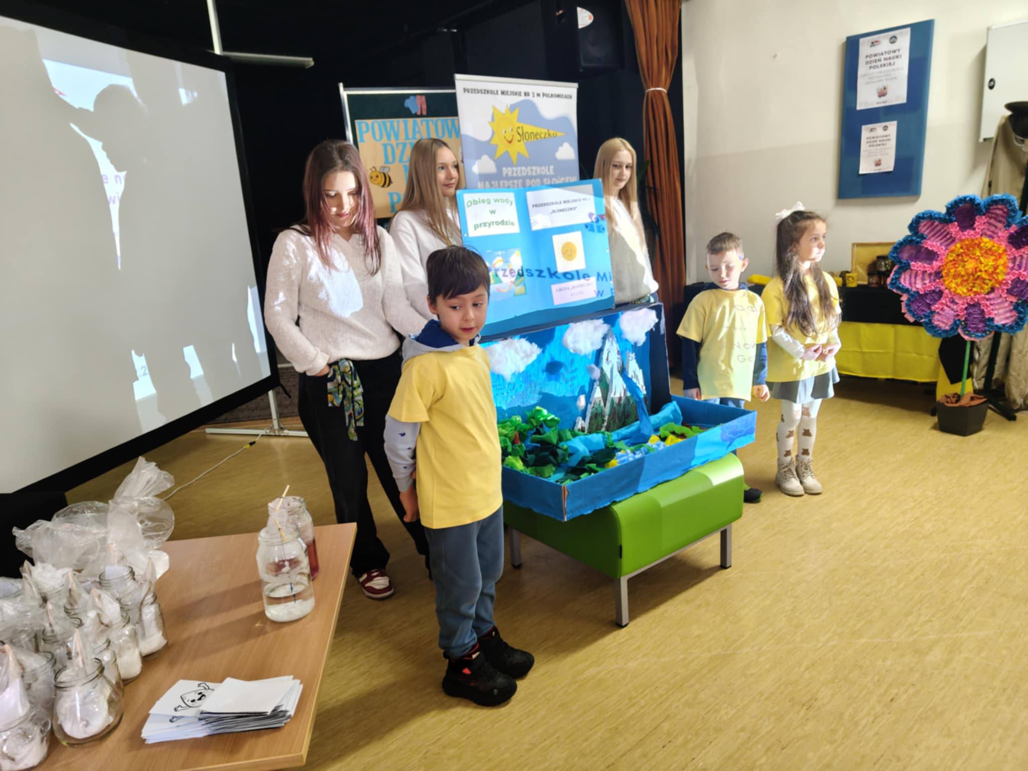 Troje dzieci ubranych w ż&oacute;łte koszulki stoi na tle makiety z obiegiem wody.