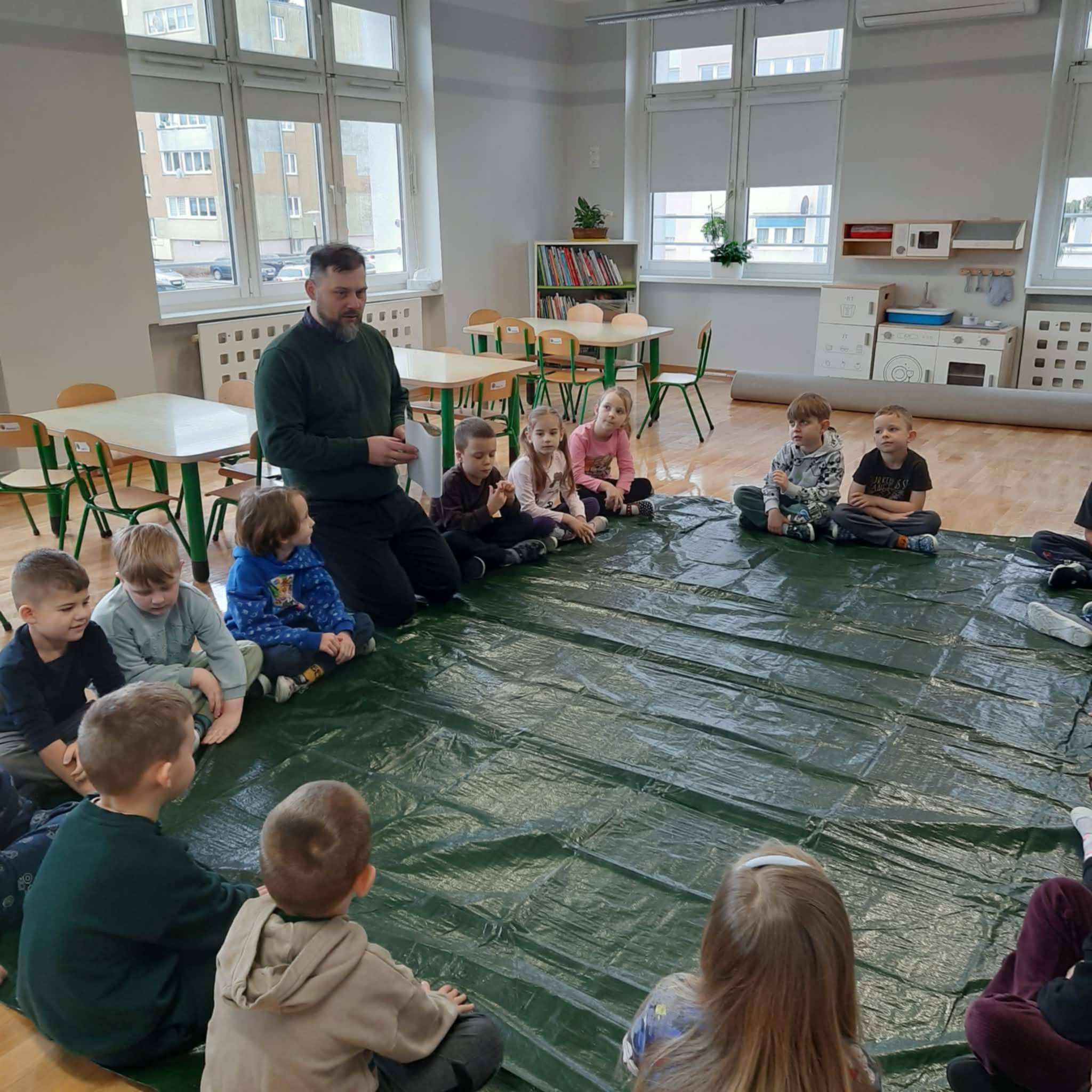 Dzieci siedzą w kole na dywanie przykrytym folią i słuchają zaproszonego gościa.