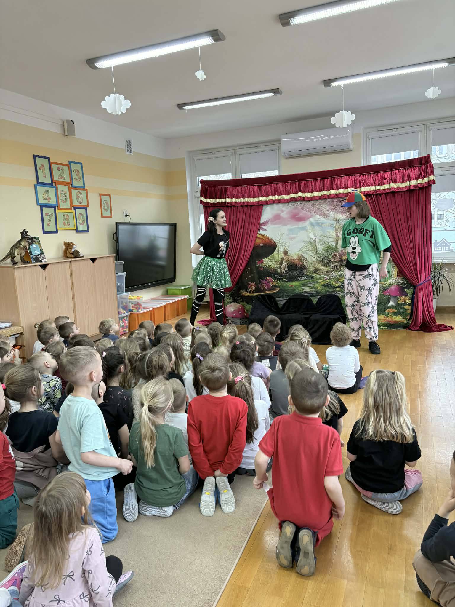 Dzieci siedzą w sali na dywanie i ogladają przedstawienie w wykonaniu dw&oacute;ch aktor&oacute;w.