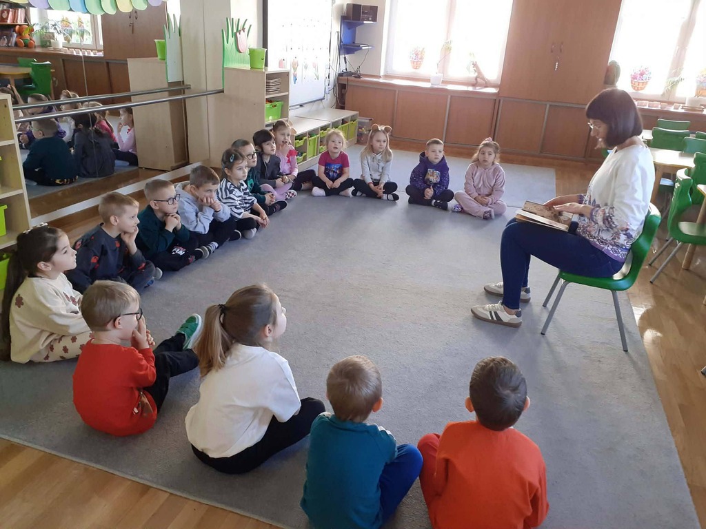 Mama jednego z dzieci siedzi na krześle i czyta dzieciom bajkę.