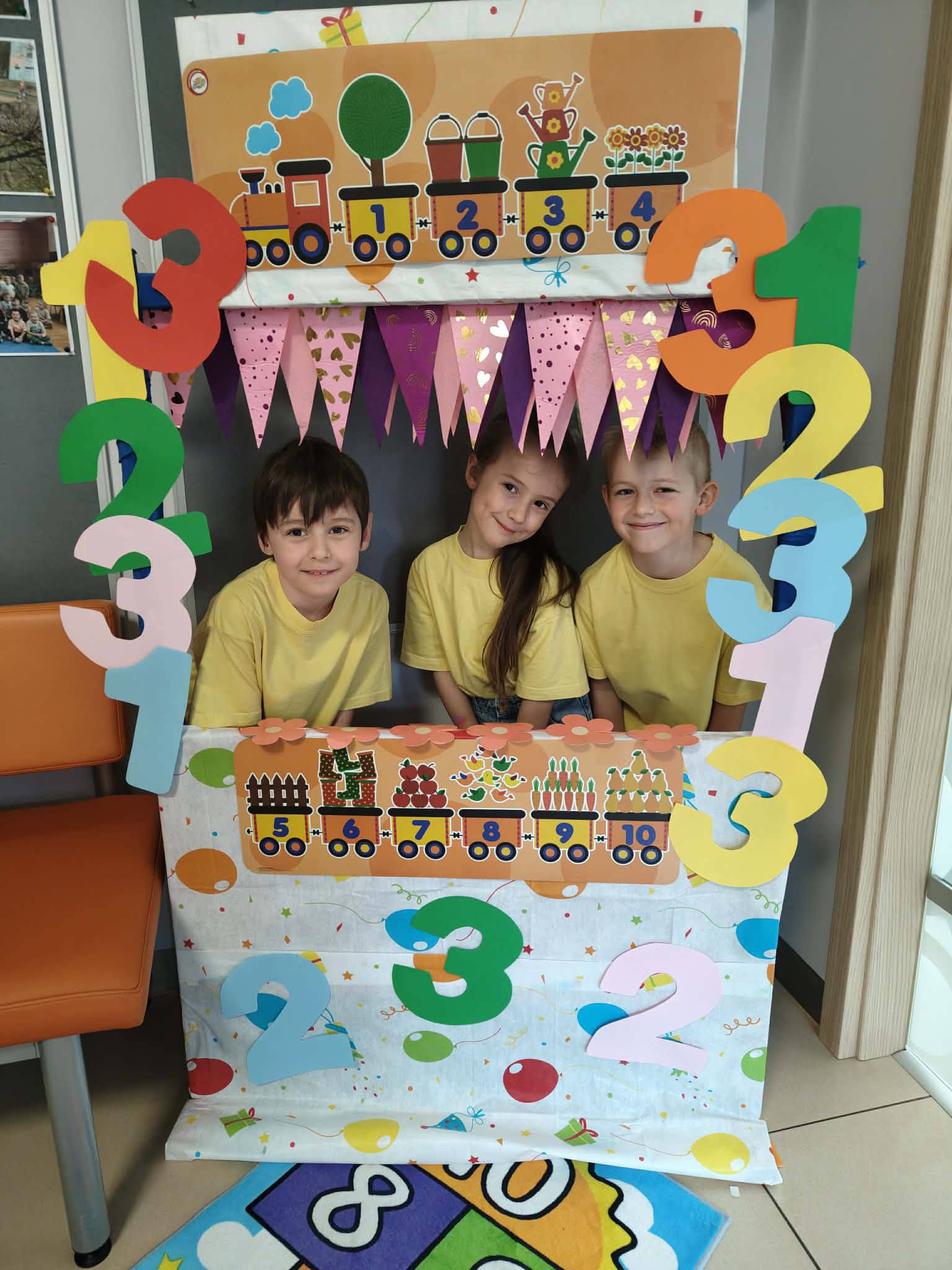 Dzieci pozują do zdjęci a w ż&oacute;łtych koszulkach stojąc w fotoramce z cyframi.