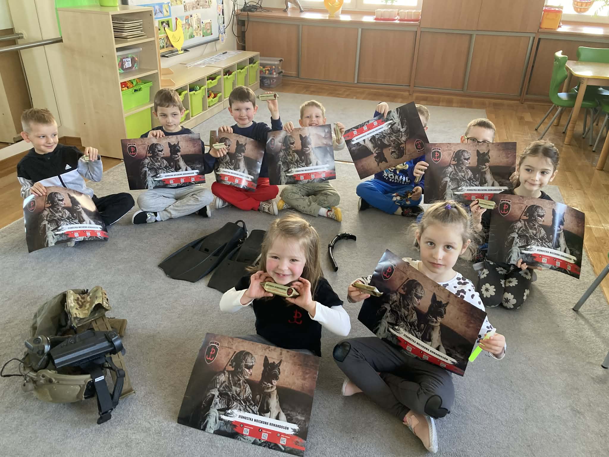 Dzieci siedzą na dywanie trzymając w rękach  przedmioty oraz obrazki związane z wojskiem.