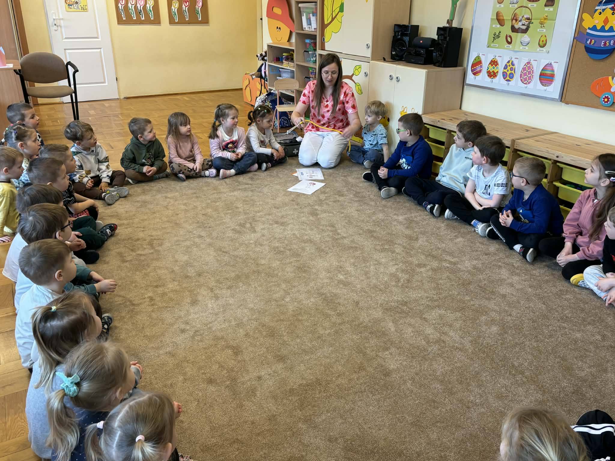 Dzieci siedzą w kole na dywanie z panią pielęgniarką.