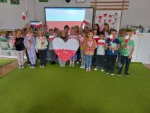 Grupa dzieci stoi trzymając wielkie serce biało- czerwone oraz flagi w ręku.