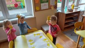 Dzieci odbijają na kartce  pomalowane żółtą farbą ręce.