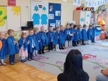 Dzieci stoją w półkolu ubrane w niebieskie pelerynki.