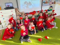 Dzieci ubrane na czerwono siedzą na dywanie i trzymają w ręku czerwone serduszka.