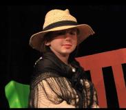 Dziewczynka w kapeluszu siedzi na krześle na scenie.