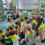 Dzieci siedzą na małych dywanikach w bibliotece i słuchają pani bibliotekarki.