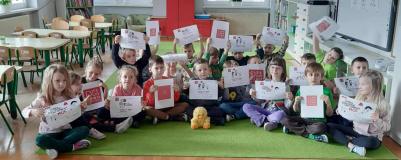 Dzieci siedzą w grupie na dywanie trzymając w rękach kartki z obrazkiem logo kampanii.